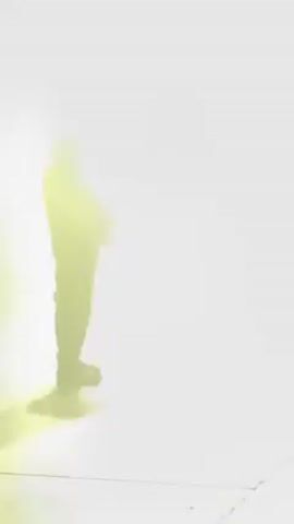 The video shows two figures in silhouette standing in a space filled with bright, diffuse light and long, horizontal streaks of light. The person on the left is heavier, wearing a jacket and dark pants with dark shoes. They are holding something in their hands and adjusting it. The person on the right is standing further back and also silhouetted. The floor has a tiled or gridded pattern. The background is almost entirely obscured by the bright light and fog or haze.