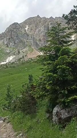 This video shows a view of a mountain landscape. In the foreground, there are green grassy slopes and some rocks. A path is visible winding through the grass. In the midground, more grassy slopes continue up the mountainside, with some scattered trees. In the background, a large, rocky mountain dominates the scene, with patches of snow visible on its upper slopes. The sky is partly cloudy.
