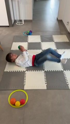 A young boy with dark hair is lying on a gray and white interlocking foam mat. He is wearing jeans, white socks, a white long-sleeved shirt, and a red belt. He starts on his stomach and uses his arms to propel himself forward while lying down, moving across the mat. The boy is carrying a green ball. There is a yellow ring near his feet with an orange, yellow, and red ball inside it. In the opposite corner of the mat is another yellow ring containing a green, dark blue, and light blue ball. The mat is on a gray tiled floor. A white cabinet and a small table stand in the background.