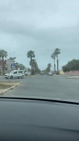 The video presents a street scene from the perspective of a vehicle moving along a road in what appears to be an urban or suburban setting, likely El Jadida, Morocco, based on the context. The road is asphalt and lined on both sides by numerous tall palm trees, suggesting a warm climate. The sky is overcast and cloudy throughout the video.

Various cars are visible on the road, both driving and parked along the shoulders. Streetlights are also present, extending into the distance on both sides of the street. At the 0:02 mark, a white van, resembling a Renault Kangoo or similar utility vehicle, passes across the screen. The van has a red and black logo on its side that clearly reads "ONCF", which stands for Office National des Chemins de Fer (Moroccan National Railways). This detail strongly indicates the location is in Morocco.

Buildings are visible behind the line of palm trees, though they are not distinctly detailed. In the latter part of the video (around 0:03-0:04), a white plastic bag can be seen briefly blowing across the road surface. The overall impression is that of a bustling street on a cloudy daytime.