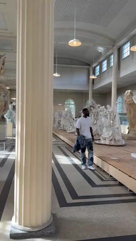 A man walks through an art museum. The man is dark-skinned and has a beard. He is wearing a white t-shirt, denim patchwork pants, and white sneakers. He carries a denim jacket over his left shoulder. He walks down the center aisle of the museum. The aisle is elevated and made of wood, with black and grey tiling on the floor on either side. There are rows of white sculptures on either side of the aisle, including various human figures, some nude. The museum has high ceilings, tall columns, and large windows. There are several modern pendant lights hanging from the ceiling. There are other people visible in the background, presumably museum visitors.