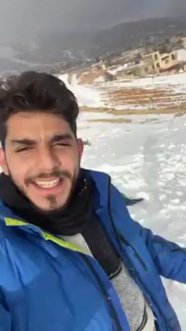 A young man with a beard and braces is smiling and taking a selfie video. He is wearing a blue jacket and a black scarf. In the background, there is a snowy landscape with a few people. Some of the people are bending over and gathering snow. There are buildings visible in the far background, and a few small trees scattered around the snowy area. The sky is overcast.