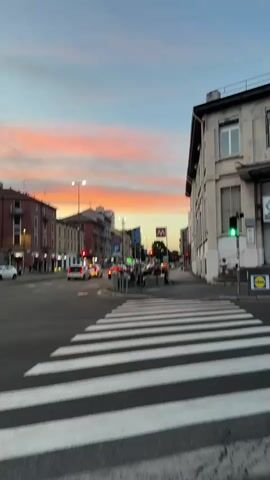 The video opens at a street intersection with a crosswalk in the foreground. In the background, buildings line the street, and a vibrant sunset with pink and orange hues illuminates the sky. Traffic lights and streetlights are visible, along with cars and a few pedestrians. A sign for the metro is also present. The scene captures the transition from day to night in an urban environment.