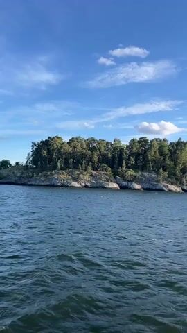 A vertical video captures a wide shot of a body of water with a small, tree-covered island in the background. The water is a deep blue with gentle waves. The island features rocky shores and dense green trees. The sky is a vibrant blue with scattered white clouds. Sunlight intermittently breaks through the clouds, creating bright reflections on the water's surface. The video appears to be taken from a moving boat.