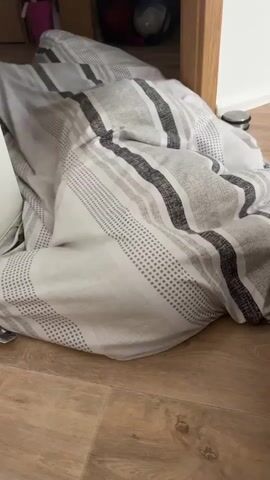 The video shows a close-up of a bed with striped bedding. The camera pans across the bed, showing the detail of the fabric and the pattern of grey and white stripes. The bedding appears to be slightly rumpled, suggesting it has been slept in or disturbed. The background is slightly out of focus but appears to be a room with wooden flooring and some furniture, possibly a wardrobe or cabinet. There is a doorframe visible on the right side of the screen. The lighting suggests an indoor environment, possibly a bedroom. No people are visible in the video.