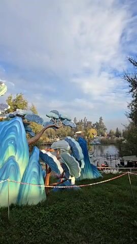 The video shows a park on a cloudy day. The foreground features a blue and white painted installation of a mountain and tree scene. A red and white barrier rope surrounds the installation.  Beyond the installation, people are walking on a path near a pond or lake.  The pond has a small structure on it and is surrounded by trees.  Further into the scene, more trees and foliage are visible.  The overall setting appears to be a botanical garden or a park decorated with art installations.