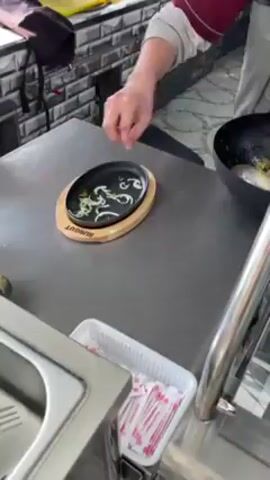 A person is cooking in what appears to be a kitchen. The cook, wearing a maroon shirt and grey vest, uses a large ladle to transfer cooked ingredients from a black wok to a sizzling plate. The sizzling plate rests on a wooden trivet with the word "BURGOUT" inscribed on it. Some raw sliced onions are already on the plate. The plate sits on a stainless steel counter. There is a white plastic container with red-wrapped chopsticks next to the sizzling plate. A sink basin and a partial view of a metal structure are visible. The background includes a tiled wall and various objects. Steam rises from the food being transferred.