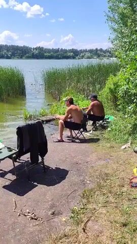 Two shirtless men are fishing by a lake on a sunny day. They are sitting on portable chairs on the shore, which is covered in dirt and some scattered debris. Both men are holding fishing rods that extend into the water. In the foreground, there is a small folding table with a green cloth and a few items on it, as well as some clothing draped over a chair. Behind the men, tall reeds grow along the water's edge, and in the distance, there is a treeline bordering the lake under a blue sky with scattered clouds. Two ducks are visible swimming in the water between the reeds.