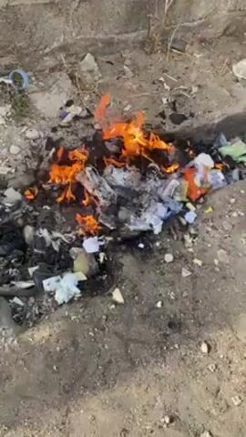 A small fire burns on the ground amongst various pieces of trash. The flames are bright orange and yellow and rise above the pile of burning debris. The ground is dirt and gravel, with larger rocks scattered around the fire. A concrete wall is visible in the background.