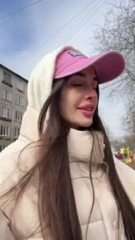 A woman is recording herself outdoors, seemingly in a residential area. She's wearing a pink baseball cap, a white hooded jacket, and has long, dark hair. Behind her is a grey, Soviet-style apartment building. She gestures towards the building and announces she is giving a weather report from Saint Petersburg, noting that the sun is even visible today. The environment suggests a cloudy or overcast day, despite her claim about the sun.