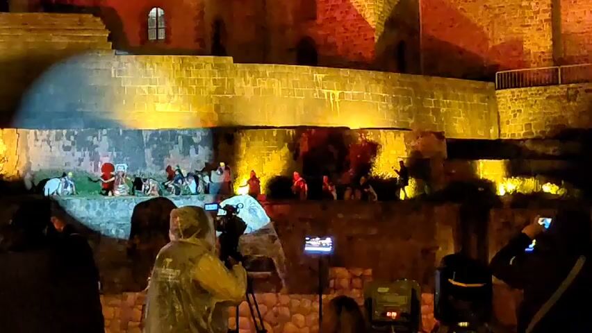At night, a live nativity scene plays out on a stone platform against the backdrop of a brick wall illuminated by red and yellow lights. Actors in costume portray the birth of Jesus. Three people stand in the foreground, facing the scene and filming or photographing it with various devices: a professional video camera, a cell phone on a tripod, and a cell phone held in hand. A spotlight shines on the nativity scene. There are more people in the background behind the camera people.