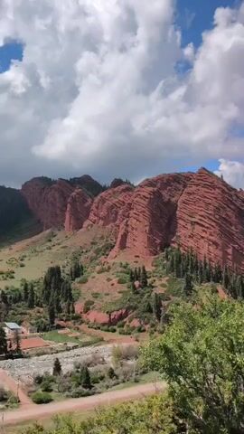 The video presents a panoramic view of a vibrant mountainous landscape under a partly cloudy blue sky. The most prominent feature is a series of rolling hills and mountains composed of striking red rock formations, characterized by distinct layered and undulating patterns. These reddish-brown slopes are partially covered with green vegetation, including numerous evergreen trees, especially in the valleys and lower sections.

As the camera pans from left to right, it reveals more of the expansive vista. Below the red rock mountains, a valley floor can be seen, with what appears to be a riverbed, some sparse vegetation, and hints of human infrastructure, possibly roads or tracks, and even what looks like a small sports court. The green foliage of trees and bushes in the foreground frames the majestic red mountains. The sunlight illuminates the landscape, highlighting the rich colors and textures of the geological formations, contrasting with the verdant greenery and the dynamic sky. The scene evokes a sense of vast natural beauty and ruggedness.
