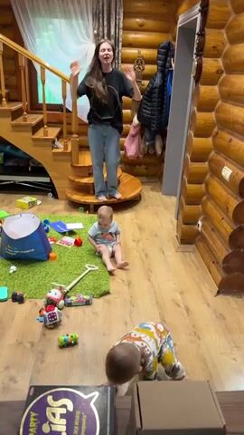 The video shows the interior of a log cabin style house. A young woman in a black t-shirt and jeans stands barefoot on a spiral staircase and does some sort of hand movements, perhaps dance-like. Two toddlers are on the floor. One is sitting on a green rug, wearing a light blue shirt and a diaper. The other is standing and walking, wearing a dinosaur-patterned onesie. Toys are scattered on the floor around the rug, including building blocks, toy cars, and a soft blue bin. There is a board game, "Alias Party" visible on the floor. The walls are made of logs, and a coat rack is visible near the staircase. A garland hangs from the ceiling.