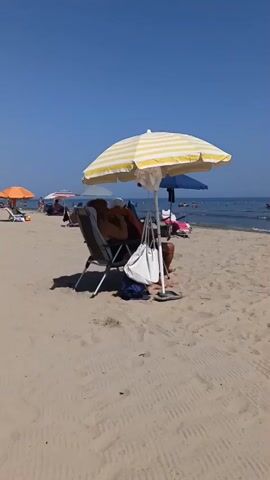 The video shows a sandy beach on a sunny day with clear blue skies and calm ocean waters. Several beach umbrellas, in various colors like yellow and white striped, orange, blue, and dark green, are set up on the sand. Beneath these umbrellas, people are seen relaxing on chairs, some appearing to be sunbathing or reading. Beach chairs and towels are visible. In the background, several people are in the ocean, some swimming and others wading. The overall atmosphere is one of leisure and recreation at a seaside resort.
