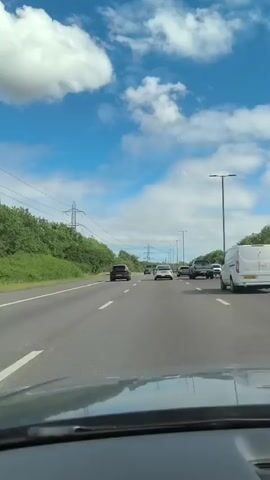 A forward-facing dashboard camera view shows a multi-lane highway on a bright, sunny day. The camera is inside a vehicle moving along the highway. To the left of the highway, there are green trees and a power line with large metal towers. The sky is blue with scattered white clouds. The highway has multiple lanes of traffic, with several cars and vans visible in front of the camera vehicle. The vehicles are moving at a steady pace. Tall streetlights line the right side of the highway.
