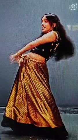 A young girl with long dark hair performs a dance in front of a gray backdrop. She is wearing a black cropped sleeveless top with a gold skirt that has a black border. The skirt has a textured pattern. She moves her arms and body fluidly, twirling and gesturing. At one point, she covers her eyes with her hand.