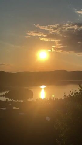 A vertical video captured from a high vantage point at sunset. The sun, a bright orb, is positioned in the upper right quadrant of the frame, casting a warm, golden glow across the scene. Wispy clouds are scattered across the sky, illuminated by the setting sun. Below, a body of water, possibly a lake or river, reflects the sunlight, creating a shimmering path. The landscape features rolling hills and mountains in the background, silhouetted against the bright sky. In the foreground, trees with green foliage frame the lower part of the image. The light suggests it is either early morning or late evening. There are no people or discernible objects in the video that would indicate specific activities or interactions. The environment is natural and scenic, dominated by the warm hues of the sunset.