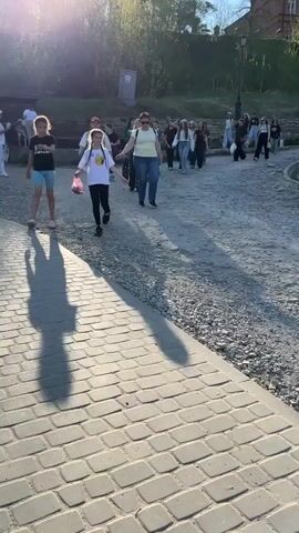 A group of people, mostly young girls, are walking on a path made of gravel and cobblestones. They are walking away from the camera towards a small hill with trees and a brick building on top. The sun is shining brightly, casting long shadows in front of the walkers. Several of the walkers are carrying bags or purses. Some of the people are smiling or talking. The environment is sunny and appears to be a park or outdoor area.