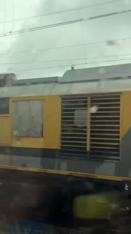 A train moves past the camera. The train cars are yellow and grey with some black stripes. The first car has a logo that appears to say "Российские железные дороги" which translates to "Russian Railways." The second car has text that says "МОДУЛЬ" which means "module." A number, 37803046, is visible on this car. The third car is grey and has a number, 37564226, at the bottom. The sky is overcast and there are water droplets on the camera lens, suggesting it is raining.