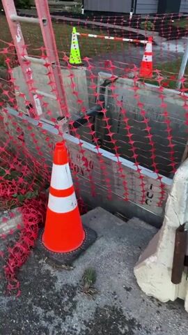 The video shows a construction site with a trench dug into the ground. The trench is shored with metal plates and wooden planks. Orange safety netting surrounds the trench. There are orange and white traffic cones placed around the site, as well as a green traffic cone in the background. A ladder is leaning against the shoring. The trench appears to be filled with muddy water. The background shows a residential street with houses. The lighting suggests it is dusk or early morning. No people are visible.