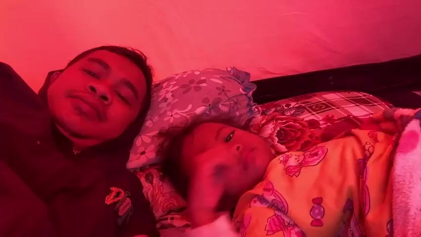The video shows two people, an adult male and a young child, lying down in what appears to be a tent. The lighting is predominantly red, creating a warm and intimate atmosphere. The adult male is in the foreground, looking directly at the camera with a slight smile. He is wearing a dark hooded jacket. To his right, the child is lying on their side, facing away from the camera and towards the adult. The child is wearing a yellow pajama set with colorful cartoon characters. They are partially covered by a pink and white blanket. There are pillows with floral patterns and a plaid blanket visible in the background. The child's hand is resting on the adult's arm or shoulder, suggesting a close relationship and interaction between them. The environment appears to be the interior of a tent, with the red fabric of the tent visible. The overall impression is one of quiet companionship and relaxation.