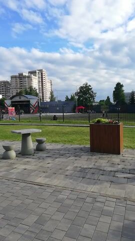 The video shows an outdoor scene with a paved walkway and a grassy area. In the foreground, there is a concrete table with two small stool-like seats. In the background, there are several apartment buildings, trees, and a fence. Some people are walking and standing around in the grassy area and on the walkway. The sky is blue with white clouds. The camera is oriented vertically.