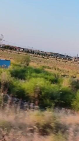 The video shows a landscape with dry grass and scattered green bushes. In the background, there are some houses and power lines. The camera is moving, suggesting it is from a vehicle. There are no people visible in the video. The sky is clear and blue. The overall impression is of an open, somewhat arid, rural or semi-urban area.