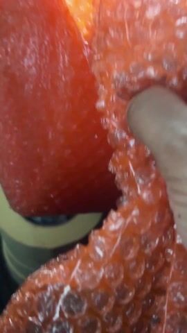 A close-up view shows a hand interacting with a large object wrapped in red-tinted bubble wrap. The main object, mostly out of focus in the upper part of the frame, appears to be a bulky, soft item, possibly a cushion or a large piece of fruit, completely encased in the translucent, reddish plastic bubble wrap. The texture of the wrapped object beneath the bubbles is indistinct but appears solid and somewhat irregular.

In the foreground, a person's right hand, with light to medium skin tone, is actively pressing down and manipulating a section of the same red bubble wrap. The bubbles in this section are clearly visible, with some appearing intact and plump, while others are distorted or have popped due to the pressure from the fingers and thumb. The act of pressing seems to be causing the bubbles to burst. The bubble wrap itself is a clear plastic with a distinct red hue, giving it a vibrant and somewhat glossy appearance under bright lighting, which creates small reflections on the individual bubbles.

The background is largely out of focus, with a blurred hint of a yellow or light green surface visible towards the bottom left, suggesting the object is resting on a surface. The overall scene focuses intensely on the tactile experience of manipulating the bubble wrap.