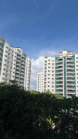 The video shows a view of apartment buildings against a blue sky with scattered clouds. Lush green trees and foliage are visible in the foreground, partially obscuring the lower portions of the buildings. The apartment buildings are tall, multi-story structures with white exteriors and balconies that feature teal-colored railings or glass. The windows are arranged in a grid pattern on the facades. The camera pans slightly to the right, revealing more of the buildings and the surrounding greenery. The overall scene suggests a residential area with an outdoor setting on a clear day.