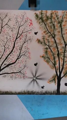 Here is a forensic description of the video:

The video shows a static shot of a painting. The painting depicts a landscape scene with two bare-branched trees, one on the left and one on the right, set against a white background. The tree on the left is adorned with red and pink foliage, while the tree on the right features green and orange leaves.

Several small, black bird silhouettes are scattered between the trees. A central design near the bottom of the painting resembles a starburst or a fan, also in black. A thin line of green and brown suggests ground level.

The painting is positioned on a surface that appears to be partially covered with a sky-blue material and possibly a white border. A black object, possibly a clamp or other tool, is visible at the top edge of the painting.