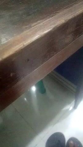 The video shows a close-up of a wooden table edge. Below the table, a tiled floor is visible, along with what appears to be the leg of a chair or table and the edge of a dark mat or rug. In the background, a green object, possibly a bottle, is dimly visible. At the bottom of the frame, the feet of a person wearing dark flip-flops are visible. The lighting suggests an indoor environment.
