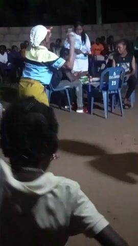 A group of people are gathered outdoors at night. In the foreground, a person wearing a light-colored shirt with a hood is facing away from the camera and appears to be walking or running. In the middle ground, a woman wearing a yellow skirt with a bird pattern and a blue and yellow shirt is dancing or moving. Behind her, a table with chairs is set up, and several people are seated. Further in the background, more people are visible, some standing and some seated. A brick wall is visible behind the seated individuals. The ground appears to be dirt or gravel. The lighting suggests it is evening or night, with artificial light illuminating the scene.