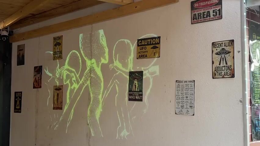 This media file shows a wall decorated with alien-themed posters and a projected image of several glowing green aliens. The aliens appear to be outlined in a wireframe style, with large heads and slender bodies. They are positioned in a dynamic way, suggesting movement or interaction.

The wall itself is white and appears to be indoors, possibly in a shed or a covered outdoor area, given the wooden beam visible at the top of the frame. Several metal signs are affixed to the wall.

One sign, top right, reads "AREA 51 TOP SECRET RESERVED FACILITY NO TRESPASSING." Another sign, to its left, says "RECENT ALIEN ABDUCTIONS IN THIS AREA" with an image of a flying saucer. Above the projected aliens, to the left, is a sign that also reads "RECENT ALIEN ABDUCTIONS IN THIS AREA." To the right of the projected aliens, there is a "CAUTION UFO ACTIVITY AREA" sign. A smaller sign below the projected aliens says "GOOD THINGS COME TO THOSE WHO WAIT." On the far right, there is a sign titled "CLASSIC UFO TYPES" with illustrations of various unidentified flying objects. There are also smaller, less legible posters or images on the wall.

The lighting in the scene is dim, with the projected aliens being the primary light source. This creates a mysterious and slightly eerie atmosphere. The overall theme is clearly extraterrestrial and UFO-related. The specific location is Nesebar, Bulgaria. No people are visible in the media file. The time of day and weather are not discernible.