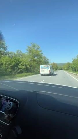 The video shows a car ride on a sunny day with a clear blue sky. The camera first shows the road ahead, then rotates to show two people in the car. A boy with short hair is singing and gesturing with his hands while sitting in the passenger seat. The girl driving the car has long brown hair and is smiling. The camera then returns to the view from the windshield, showing the road with green trees on both sides and a white van driving ahead.