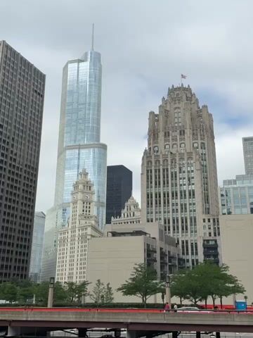 The video shows a wide shot of the Chicago skyline, featuring several tall buildings. In the foreground, a red bridge with railing is visible, with a few green trees and some people walking on the path above it. A river runs beneath the bridge, with several barges or boats docked or passing by. The buildings in the background include a prominent glass skyscraper with a spire, an ornate, gothic-style building, and a lighter-colored, more traditional skyscraper with a clock tower. The sky is overcast, suggesting a cloudy day. The camera is positioned to capture the impressive scale of the urban architecture.