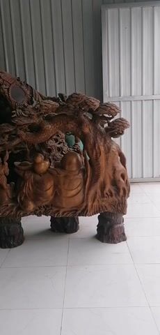 The video showcases a collection of intricately carved wooden sculptures, set against a backdrop of a gray, corrugated metal wall. A large sculpture depicting figures beneath a tree is shown in the foreground. The camera pans across the room to reveal other wooden carvings, including a chair-like structure with large holes, a statue of a smiling Buddha holding flowers, and various abstract tree-like forms. The floor is tiled with white square tiles.