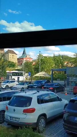 The video shows a view through a window, likely from a vehicle, looking out at a street scene with a castle in the background. In the foreground, there are several parked cars, including white SUVs and a dark colored SUV. There is also a red van. A person wearing a high-visibility vest and holding a pole is standing near a bus. The bus is white and appears to be stopped, with several people visible inside. There are also several traffic signs, including a parking sign and a sign with a picture of a bus and a car with a red circle and slash through it, indicating that buses and cars are prohibited. The sky is blue with some clouds. The environment appears to be a town or city, with buildings and trees visible. The overall scene suggests a tourist area, possibly a parking lot or a bus stop near a historical landmark.