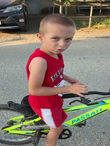 A young boy with blonde hair and blue eyes is sitting on a neon green bicycle. He is wearing a red sleeveless t-shirt with a white graphic and red shorts with white stripes. The boy is looking at the camera and smiling. The bicycle has the word "PASSATI" printed on the frame. The background shows a paved surface, a car, and some buildings.