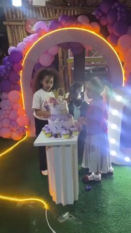 At 00:00, a birthday party is underway. A three-tiered purple cake, decorated with butterflies and flowers, sits on a white pedestal. A young girl with curly brown hair, wearing a white t-shirt with a graphic, is about to cut the cake. To her right, a young boy in a plaid shirt and a young girl in a red cardigan and a polka-dot skirt watch intently. The scene is illuminated by a large, illuminated archway of purple and pink balloons and a string of warm, yellow lights. A fluffy, white number 10 is placed on top of the cake, and the name "Tamara" is written in gold icing. The setting appears to be outdoors, with artificial grass on the ground and a wooden structure in the background, suggesting a festive gathering. The lighting is dim and atmospheric.