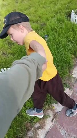 The media file is a first-person perspective video clip featuring a young child running outdoors. The child, wearing a yellow t-shirt with a black graphic and a black baseball cap with a yellow Batman logo, is being held by the hand and pulled along by the unseen videographer. The child is wearing dark brown pants and grey sneakers. The setting is a grassy area with a stone pathway. The scene appears to be during daylight, with natural lighting. The child's body language suggests movement and possibly playfulness. There is no visible text in the video, apart from the graphic on the child's shirt. The location is identified as Medvedevka, Russia. The weather appears to be mild and possibly sunny, given the bright lighting and green vegetation. The overall mood seems to be lighthearted and casual, capturing a moment of a child playing outdoors.
