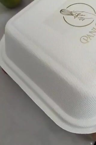 A close-up shot focuses on a white, rectangular takeout container with a textured lid. The container appears to be made of a biodegradable material. A circular logo with a drawing of a whisk and text is visible on the lid, along with the partially obscured text "Pani Cakef". A hint of red is visible beneath the container's rim. The background is blurred, suggesting a neutral surface. The lighting appears to be from an overhead source, casting a soft glow on the container. The overall impression is one of food packaging ready for serving or transport. No people are visible, and the setting is ambiguous due to the close-up framing. The image does not provide information about the time of day, weather, or specific emotions.