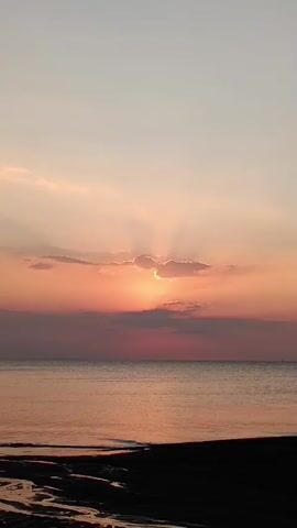 A time-lapse video of a sunset over the ocean. The sun is partially obscured by clouds, casting rays of light across the sky. The water is calm, and the beach in the foreground is dark. The colors of the sky range from soft pinks and oranges to a pale grey.