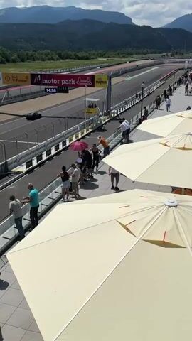 A race car drives on a track with spectators watching from above. The track is lined with barriers and advertising signs. The spectators are standing on a balcony area with several large umbrellas. The weather appears to be sunny with some clouds. Mountains are visible in the background.