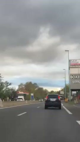 The video shows a car driving on a highway under a cloudy sky. A black SUV is in front of the camera car. There are other cars on the road and a gas station and commercial buildings are visible on the right side of the road. Streetlights line the side of the road. The overall scene appears to be suburban.