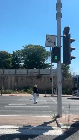 A lone pedestrian, wearing a yellow hat and dark clothing, walks across a crosswalk on a sunny day. The pedestrian is seen from behind, carrying a white bag over their shoulder. The crosswalk is clearly marked with white stripes on a dark asphalt road. In the background, a brick wall topped with a fence runs horizontally, with lush green trees visible above it. A traffic light pole stands prominently in the foreground, displaying a green pedestrian signal and a green vehicle light. Above the pedestrian signal, a sign with an image of a hand pointing down and a button icon is affixed to the pole, indicating a push-button for traffic signals. The scene appears to be in Azor, Israel, based on the provided location context. The bright sunlight suggests it is daytime, and the clear blue sky indicates fair weather. A white scooter is partially visible in the lower right corner of the frame, suggesting a moderately urban or suburban environment. The overall atmosphere is one of ordinary daily life.