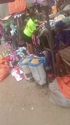 The video depicts a busy outdoor market scene. A woman in a bright green shirt and black shorts is rummaging through a pile of clothes on a table. Next to her are two large sacks filled with a yellow substance, possibly grain or flour. A person in a blue patterned shirt and shorts is sitting on a wooden structure nearby. In a different area of the market, a woman is sitting on the ground holding a baby, while a child stands beside her. There are more sacks in this area, along with a mat covered in what appear to be small, green objects. Clothing items are hung on hangers overhead. The market is bustling with activity, as suggested by the presence of numerous vendors and shoppers.