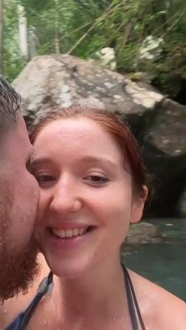 A couple is taking a selfie video in front of a cascading waterfall in a tropical environment. The woman, who is redheaded and fair-skinned, is smiling at the camera. The man, who is bearded and also fair-skinned, is holding the camera close, his face pressed near hers. Both of their faces are wet. Behind them, a multi-tiered waterfall descends over dark rocks, surrounded by lush green vegetation. Other people can be seen swimming in the pool beneath the waterfall.