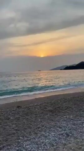 The video starts with a view of a beach at sunset. Waves lap the shore, and there are mountains in the distance. The camera then pivots to reveal a woman in a gray puffer vest standing on the beach, looking at the camera. Behind her, there's a steep, verdant hillside.