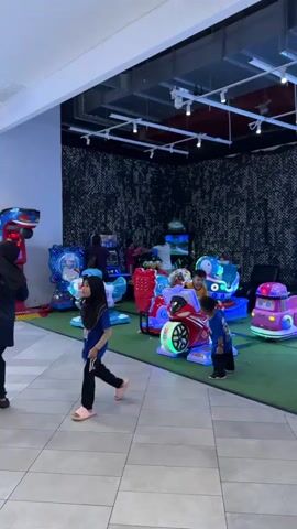 The video shows an indoor arcade with several children playing on coin-operated amusement rides and arcade games. In the foreground, a young boy in a blue shirt walks from right to left. To his right, a red motorcycle-shaped ride has a child sitting on it. Further back, a blue airplane-shaped ride with a star decal is visible, and beyond that, a Hello Kitty themed ride. In the background, several adults and children are gathered around arcade game machines, with some appearing to play. The setting appears to be a well-lit indoor space, likely a family entertainment center, with a black and white patterned wall in the background and fluorescent lighting overhead. The floor transitions from tiled to a green artificial turf area where the rides are located. The overall atmosphere seems lively and fun, with the brightly colored rides and the children's engagement suggesting a positive and active environment. There is no indication of the time of day or weather conditions. The location context suggests this is likely in Ampang Jaya Municipal Council, Malaysia. There is text on the blue airplane ride that appears to read "Princess" or a similar word.