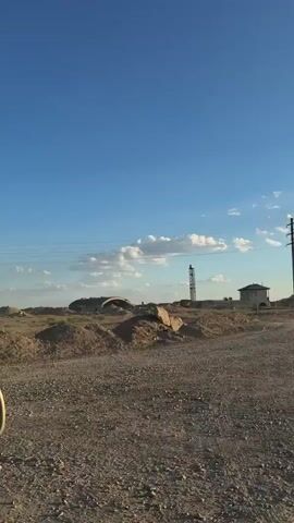 The video shows a dry, barren landscape under a clear blue sky. The ground is covered in dirt, rocks, and sparse, dry vegetation. In the distance, there are some dilapidated structures, possibly abandoned buildings or industrial facilities, along with power lines stretching across the scene. The camera appears to be mounted in a vehicle, as indicated by the side mirror visible in the lower left corner of the frame. The overall impression is one of an arid, undeveloped, or post-industrial environment.
